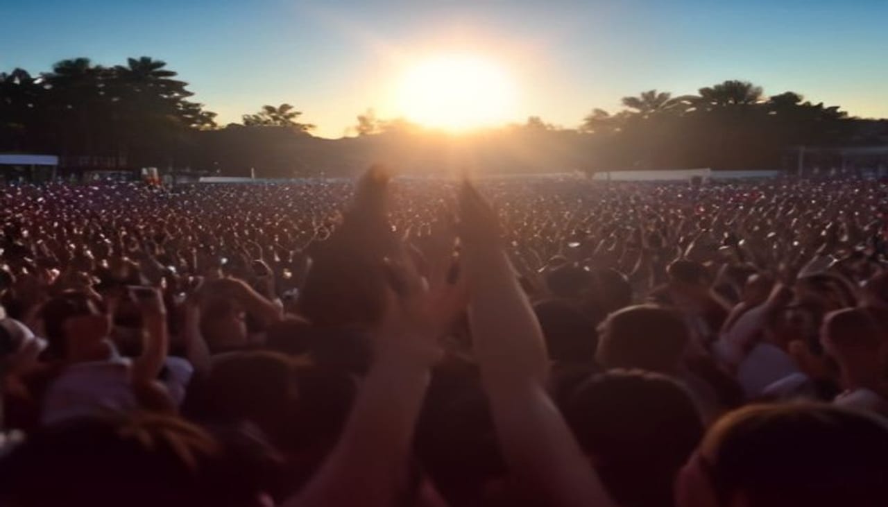 A big crowd seen from behind at an open-air party, arms raised towards a low sun setting behind palm trees